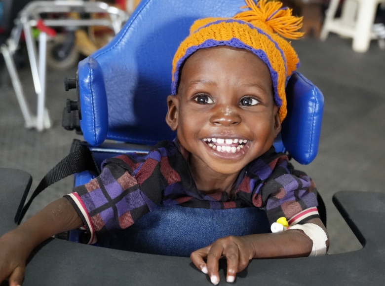 A child wearing a hat sitting in a wheelchair smiling.