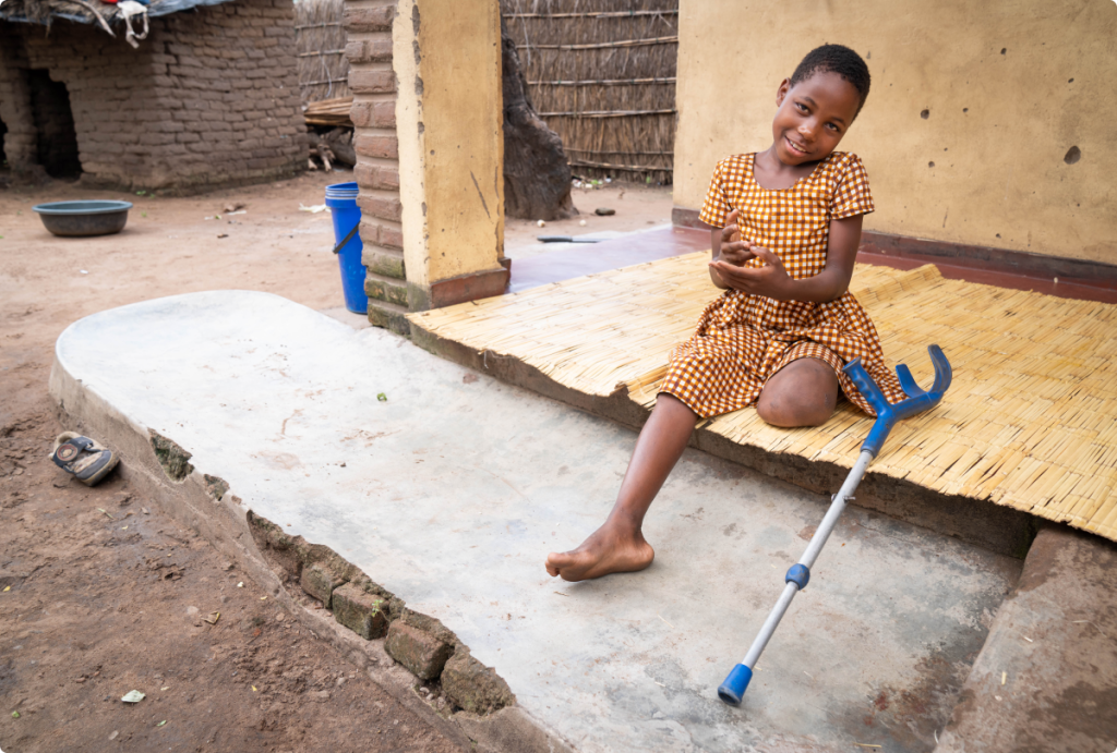A smiling girl with a leg amputation sits outside a house with her assistive device.