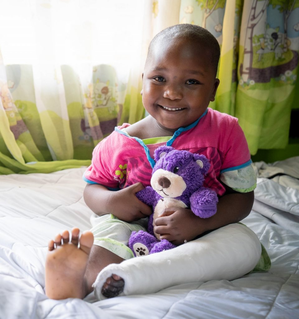 A smiling girl holds a purple teddy bear while sitting in a hospital bed with a cast on her right leg.