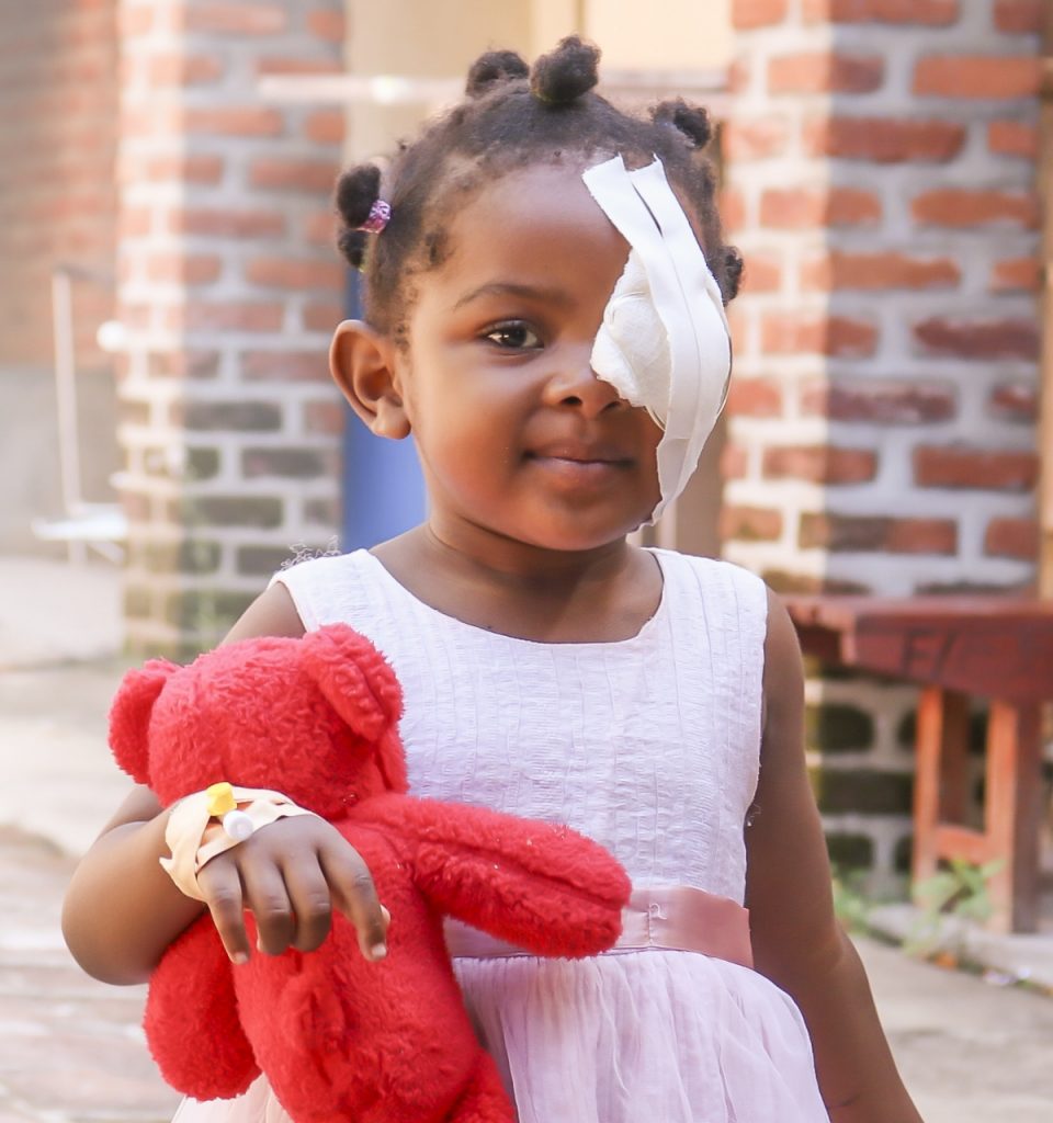 Young girl slightly smiling with eyepatch over left eye while holding a teddy bear.