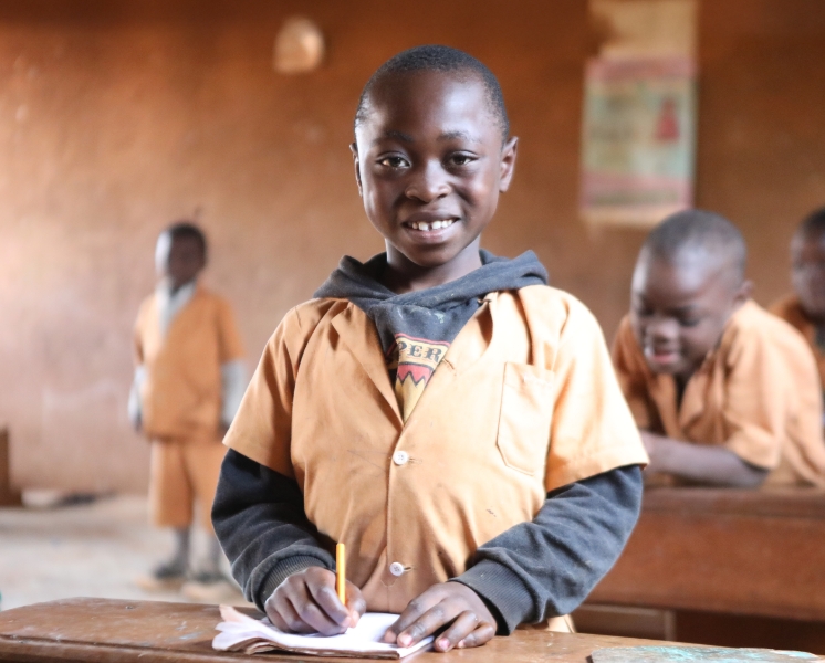 A smiling boy standing behind a desk in a classroom with a pencil and notebook.