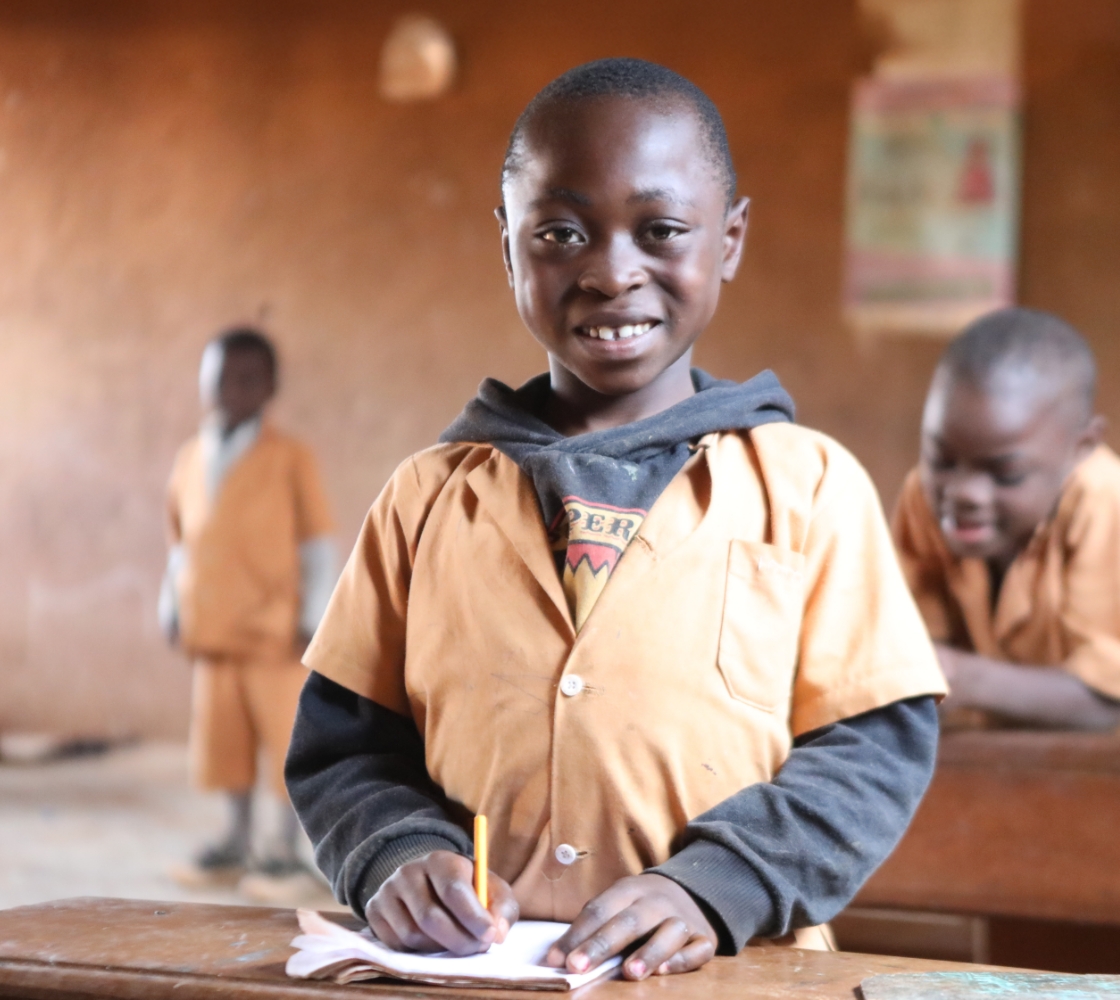 A smiling boy standing behind a desk in a classroom with a pencil and notebook.