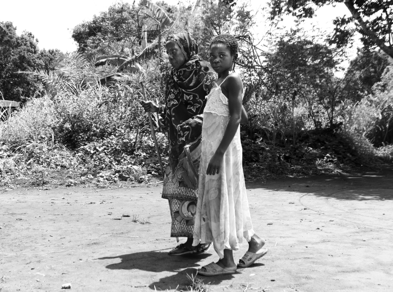 Serious child leading grandmother who is blind across uneven ground outside.