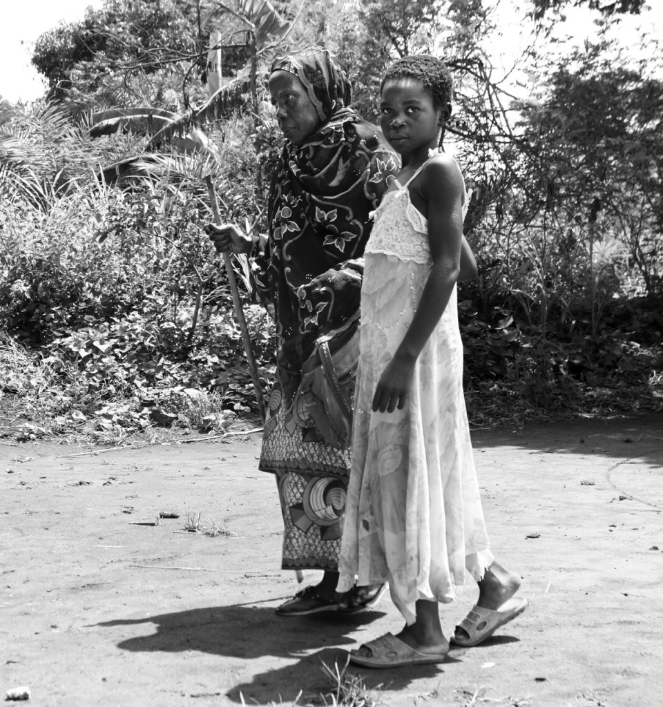 Serious child leading grandmother who is blind across uneven ground outside.