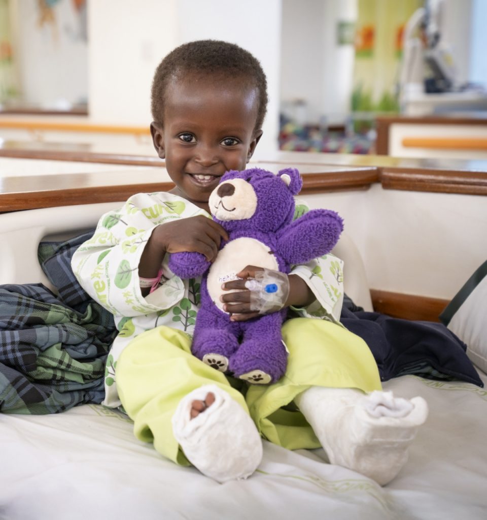 A smiling young boy sitting on a hospital bed with casts on both legs, holding a purple teddy bear.